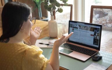 Woman in a yellow shirt gesturing at a laptop screen during a video call at a wooden table, with a notebook and a vase nearby, conveying engagement.